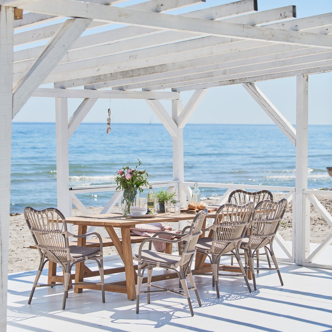 White wooden pergola with a beach view, surrounded by natural wicker chairs and a wooden dining table on a light surface.