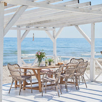 White wooden pergola with a beach view, surrounded by natural wicker chairs and a wooden dining table on a light surface.