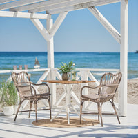 Beachside dining set featuring beige wicker chairs and a light wood tabletop, surrounded by white structure and blue ocean.