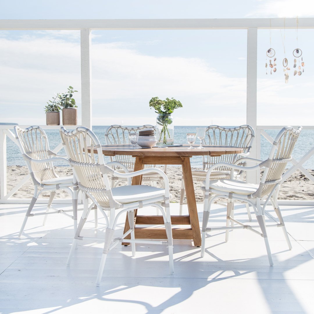White wicker chairs around a natural wood table, set against a beach backdrop with soft blue skies and sunlight.
