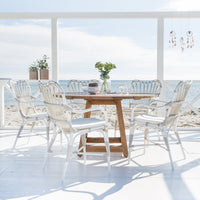 White wicker chairs around a natural wood table, set against a beach backdrop with soft blue skies and sunlight.