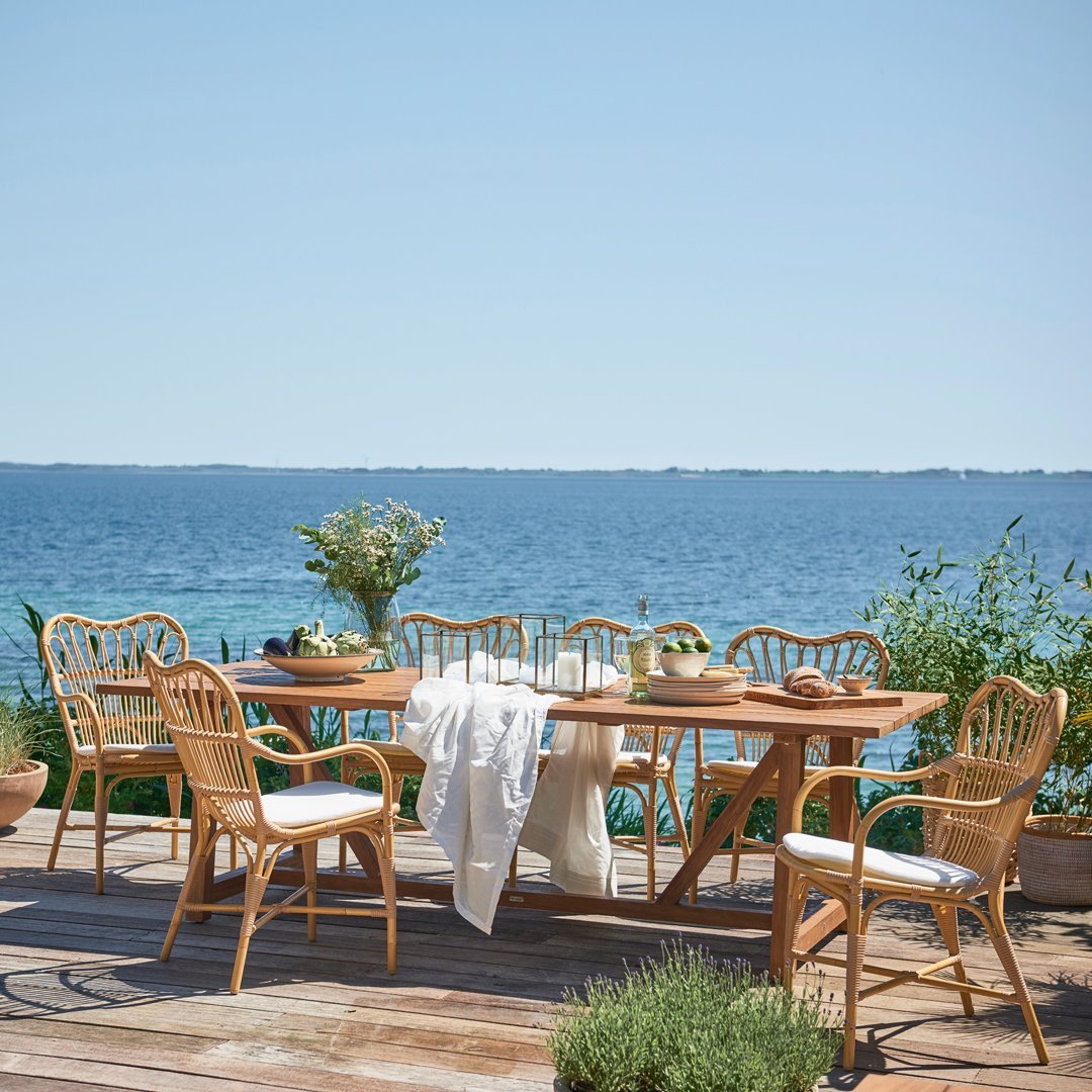 Natural wood table and wicker chairs in light tones with white cushions, set against a serene blue water backdrop.