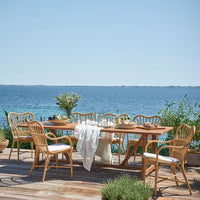 Natural wood table and wicker chairs in light tones with white cushions, set against a serene blue water backdrop.