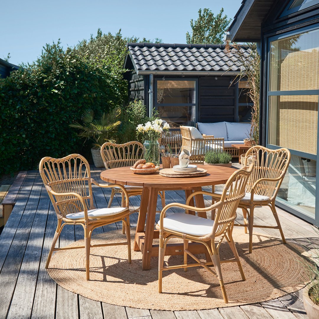 Rattan dining set with natural wood tones and white cushions, surrounded by greenery and a beige round rug.