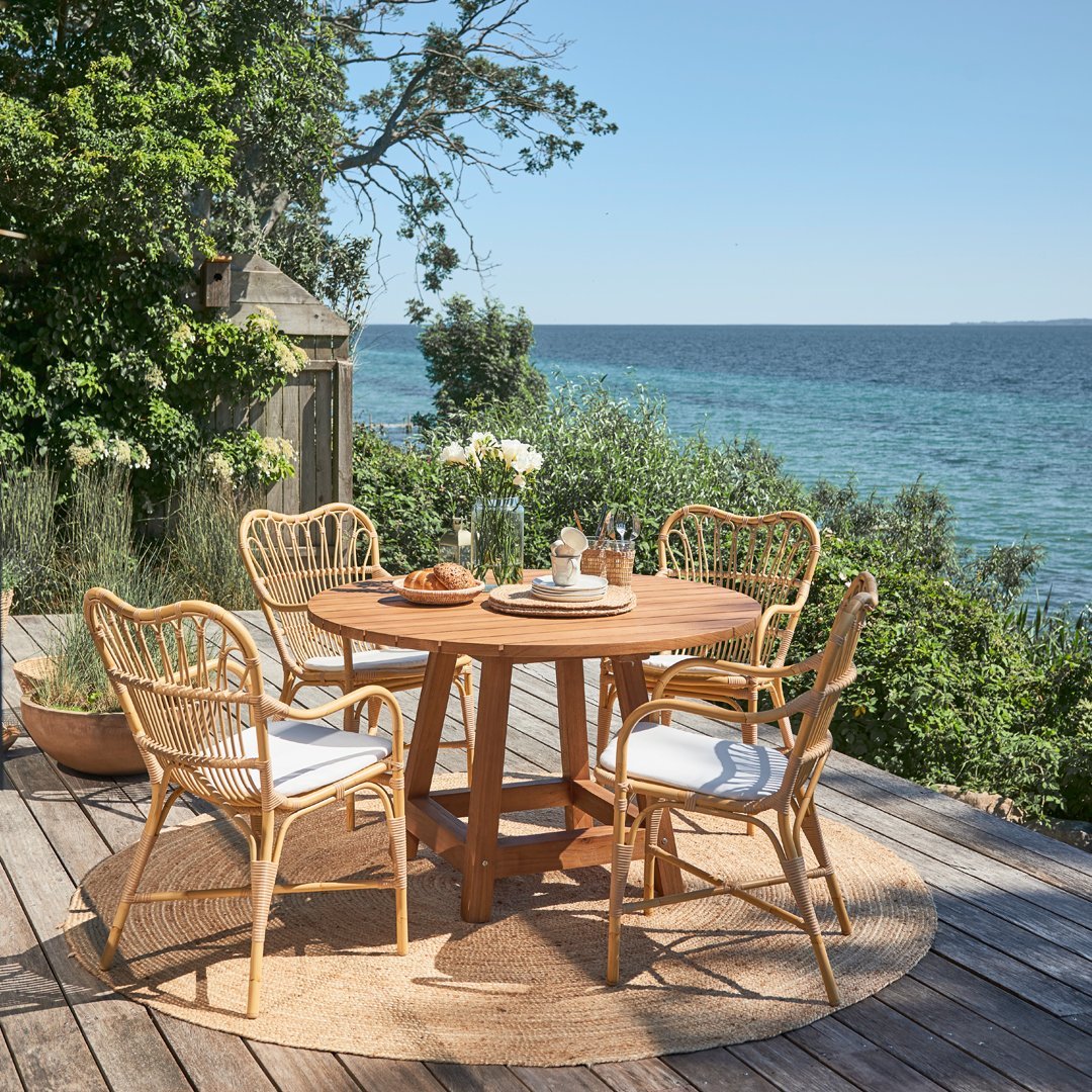 Natural wood table with rattan chairs, featuring white cushions and a woven round rug, surrounded by greenery and ocean view.