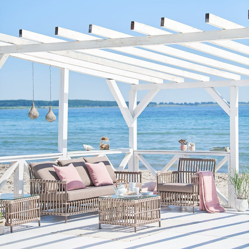 Cozy outdoor seating in muted pinks and earthy tones, set against a bright blue ocean backdrop. Relaxing coastal decor.