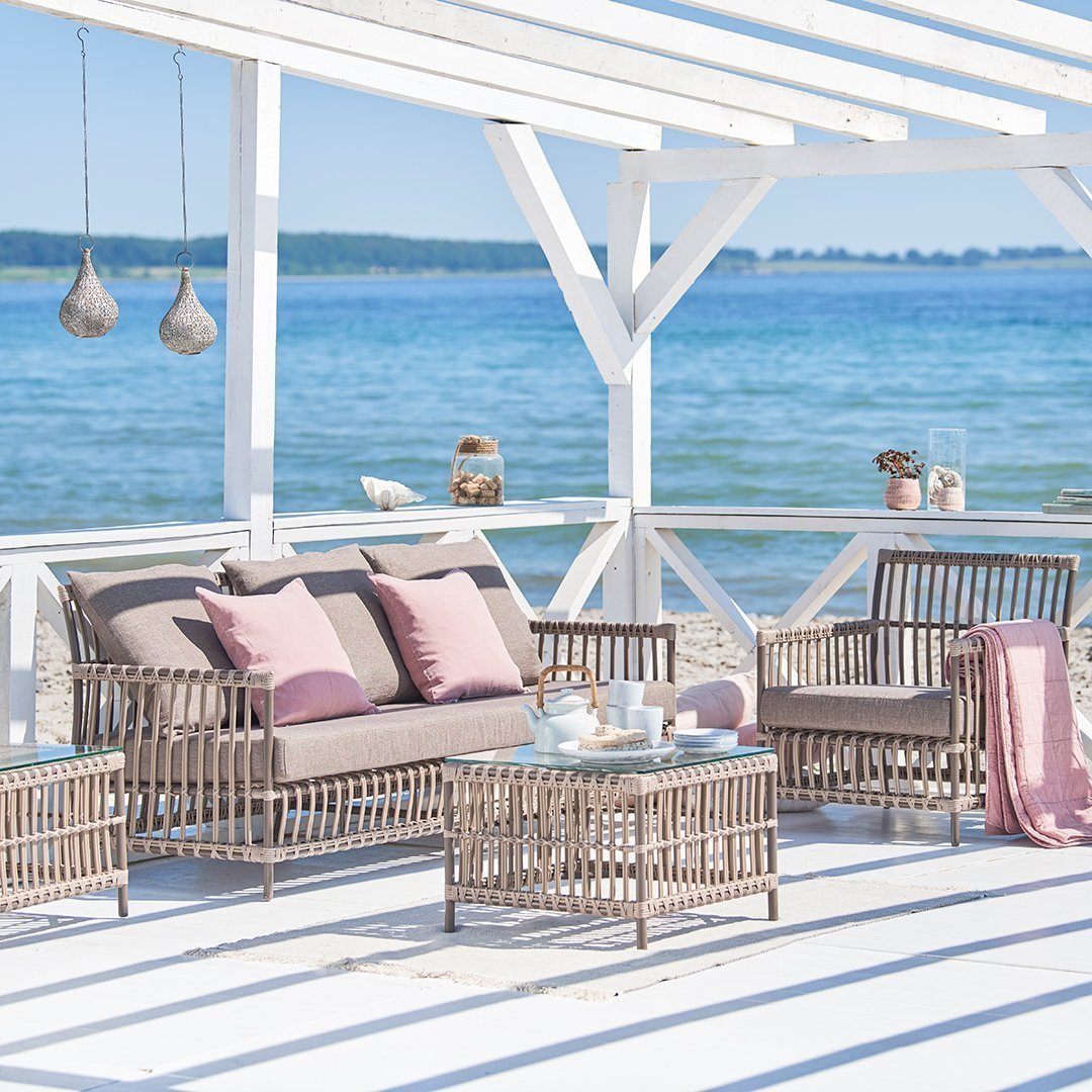 Rattan outdoor furniture set in neutral tones with pink cushions, beachside ambiance, and blue water backdrop.