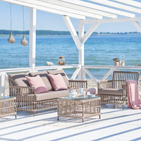Rattan outdoor furniture set in neutral tones with pink cushions, beachside ambiance, and blue water backdrop.