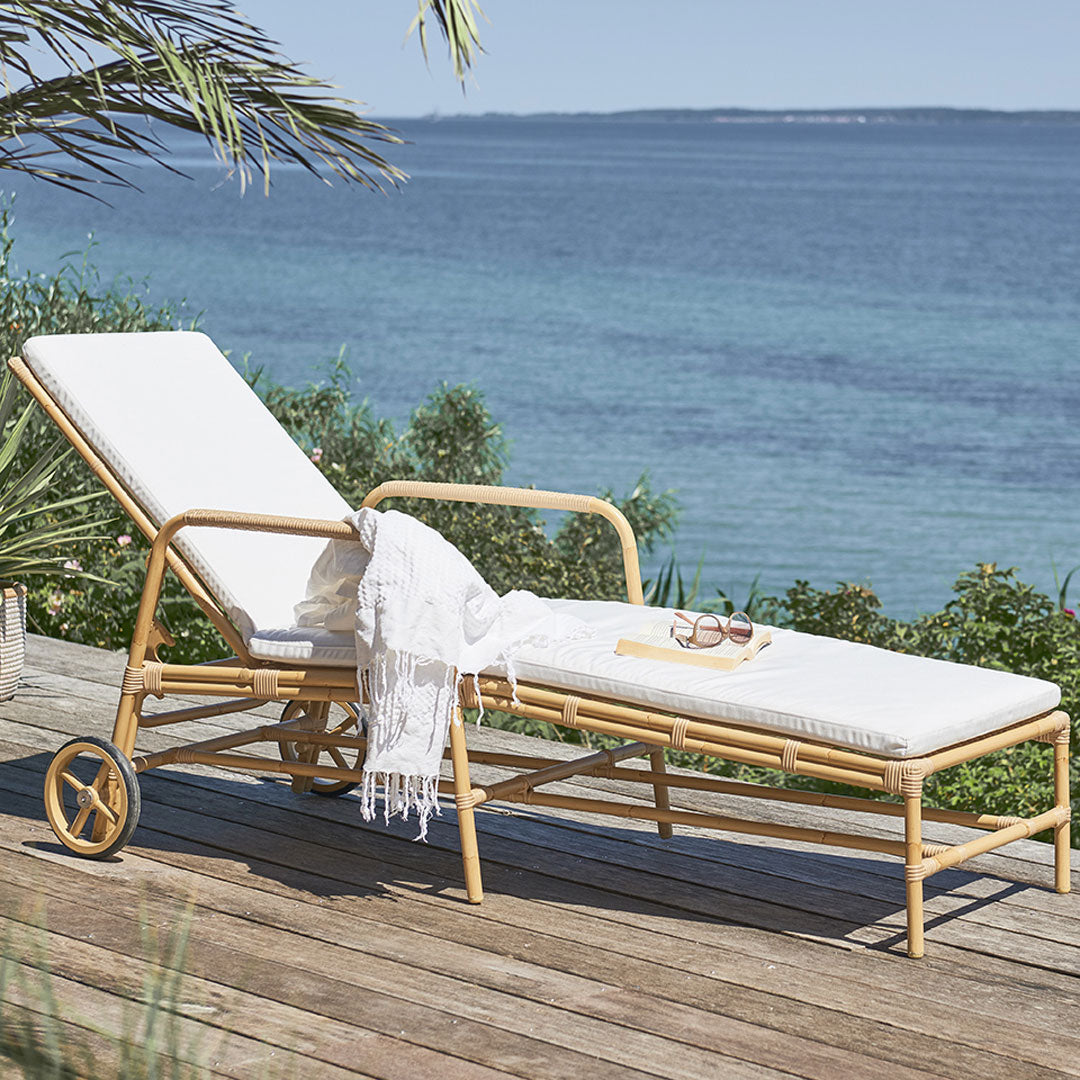 White cushioned lounge chair with natural rattan frame, set against a serene blue ocean backdrop. 