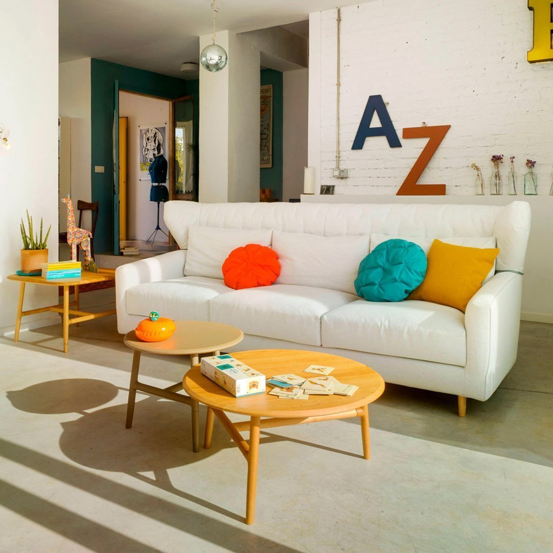 Bright white sofa with colorful cushions in orange, turquoise, and yellow; complemented by wooden coffee tables against a modern backdrop.