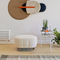 Round cream ottoman on light wood floor, paired with woven decor in beige and navy accents. Modern, minimalist design.