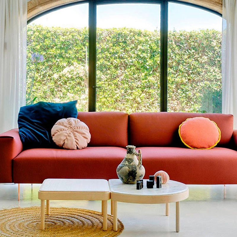 "Mid-century modern sofa in rust red with blue and pink cushions, set against bright green outdoor views."