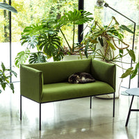 Green upholstered sofa with minimalist design, set against a backdrop of lush greenery and natural light.