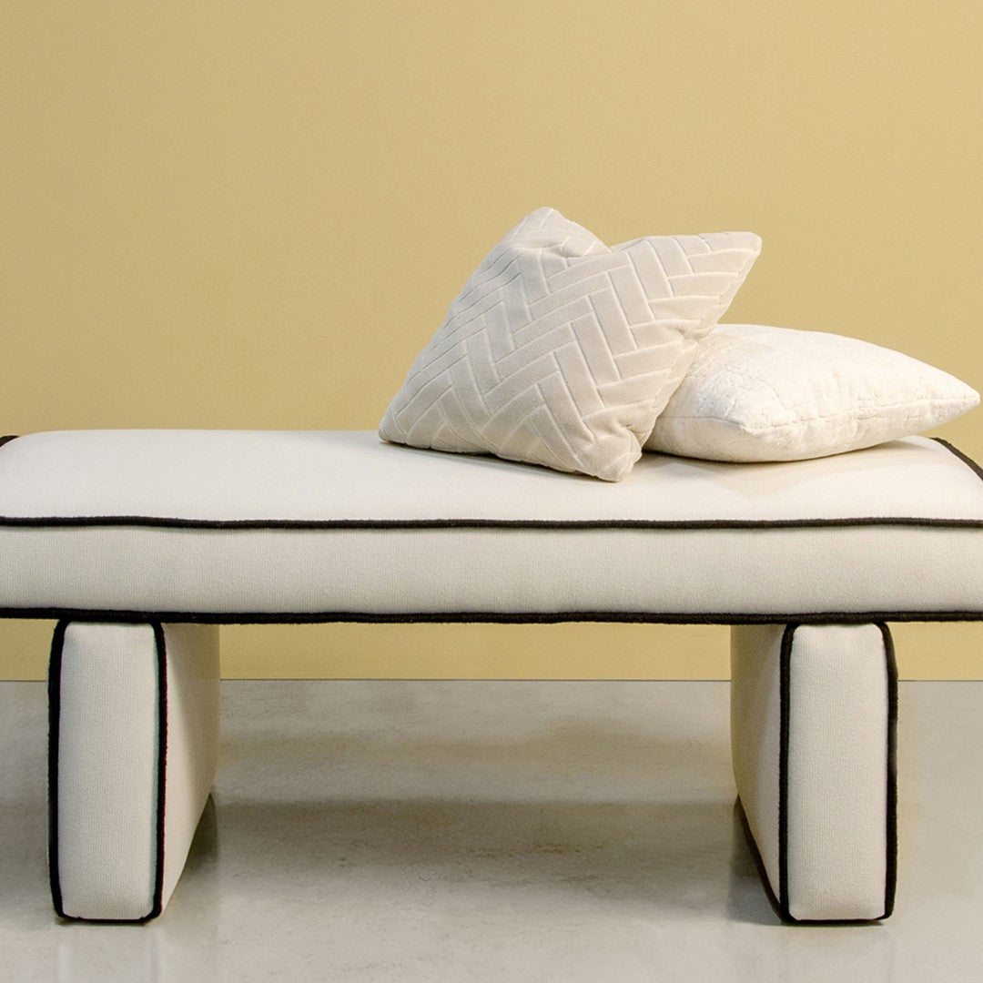 White bench with black piping, featuring textured cream and white pillows against a soft yellow background.