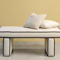 White bench with black piping, featuring textured cream and white pillows against a soft yellow background.