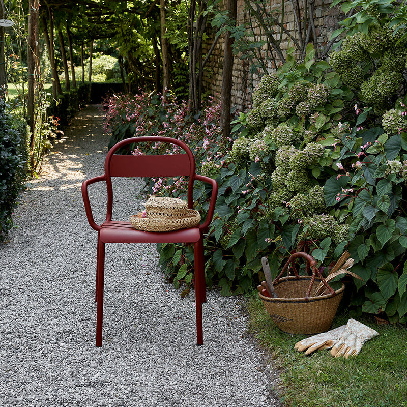 Red metal chair surrounded by lush green foliage and blooming flowers, set on a gravel pathway.