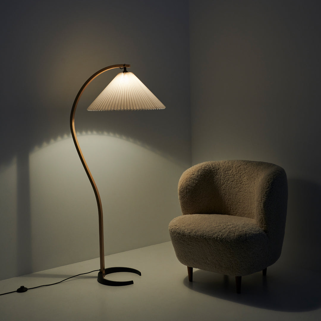 Stylish gold floor lamp with a woven white shade, complemented by a textured cream armchair in a minimalist setting.