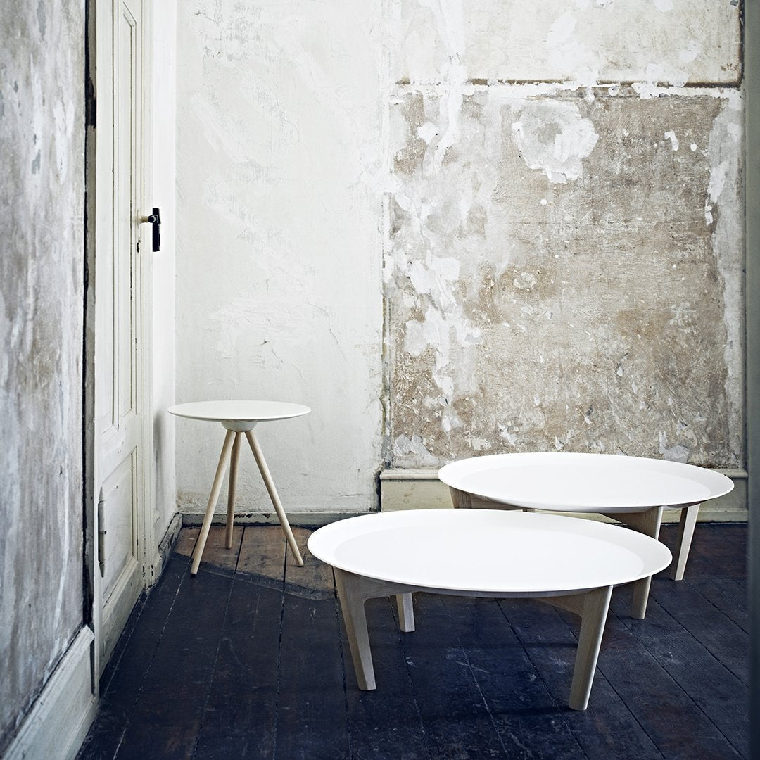 Three minimalist round coffee tables with white tops and natural wood legs, set against a textured wall.