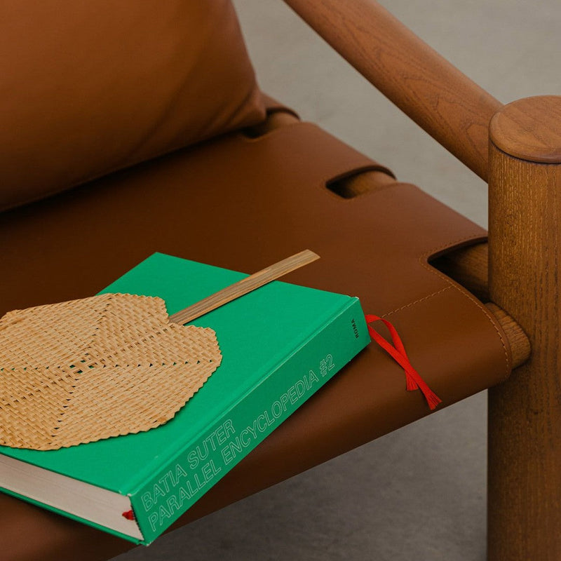 Brown leather chair with a green book and a light brown woven fan. Modern design with warm, earthy tones.