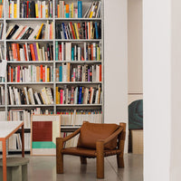 "Brown leather chair with wooden frame in modern setting, surrounded by colorful book spines on a backdrop of white walls."