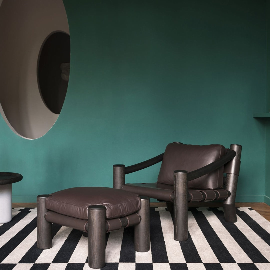 Brown leather chair with matching ottoman on bold black and white striped rug against a teal wall. Modern design elements.