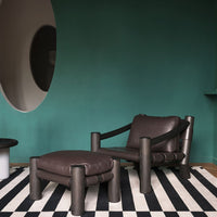 Brown leather chair with matching ottoman on bold black and white striped rug against a teal wall. Modern design elements.