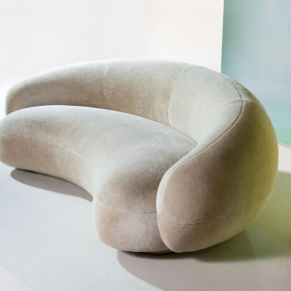 Curved, beige velvet sofa with a smooth, minimalist design and elegant silhouette on a clean background.