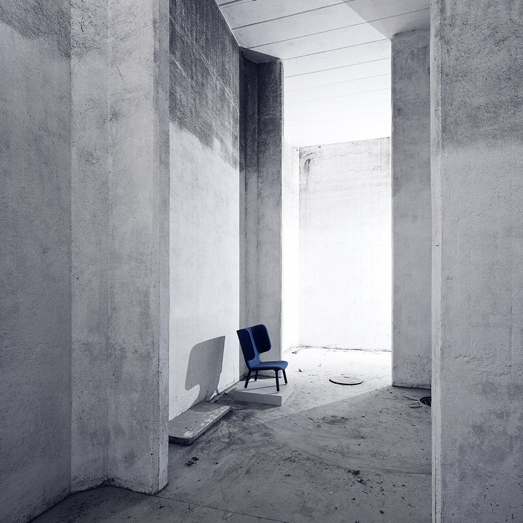 Minimalist design featuring a blue chair in a stark, concrete space with gray walls and soft shadows.