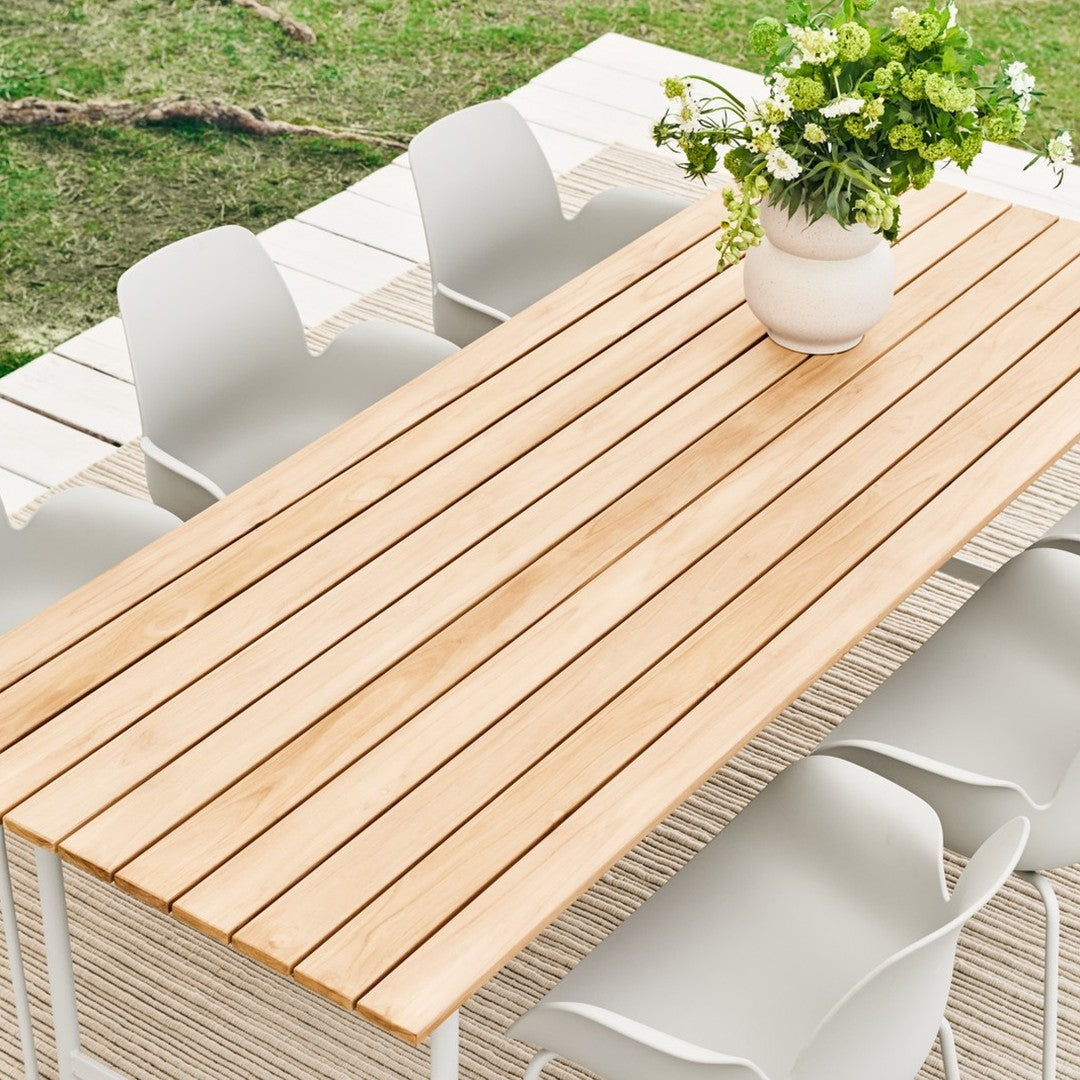 Light wood slatted dining table with six white minimalist chairs, featuring a fresh flower arrangement in a vase.