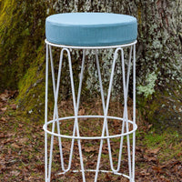 Light blue upholstered seat on a white geometric metal bar stool, set against a natural background.