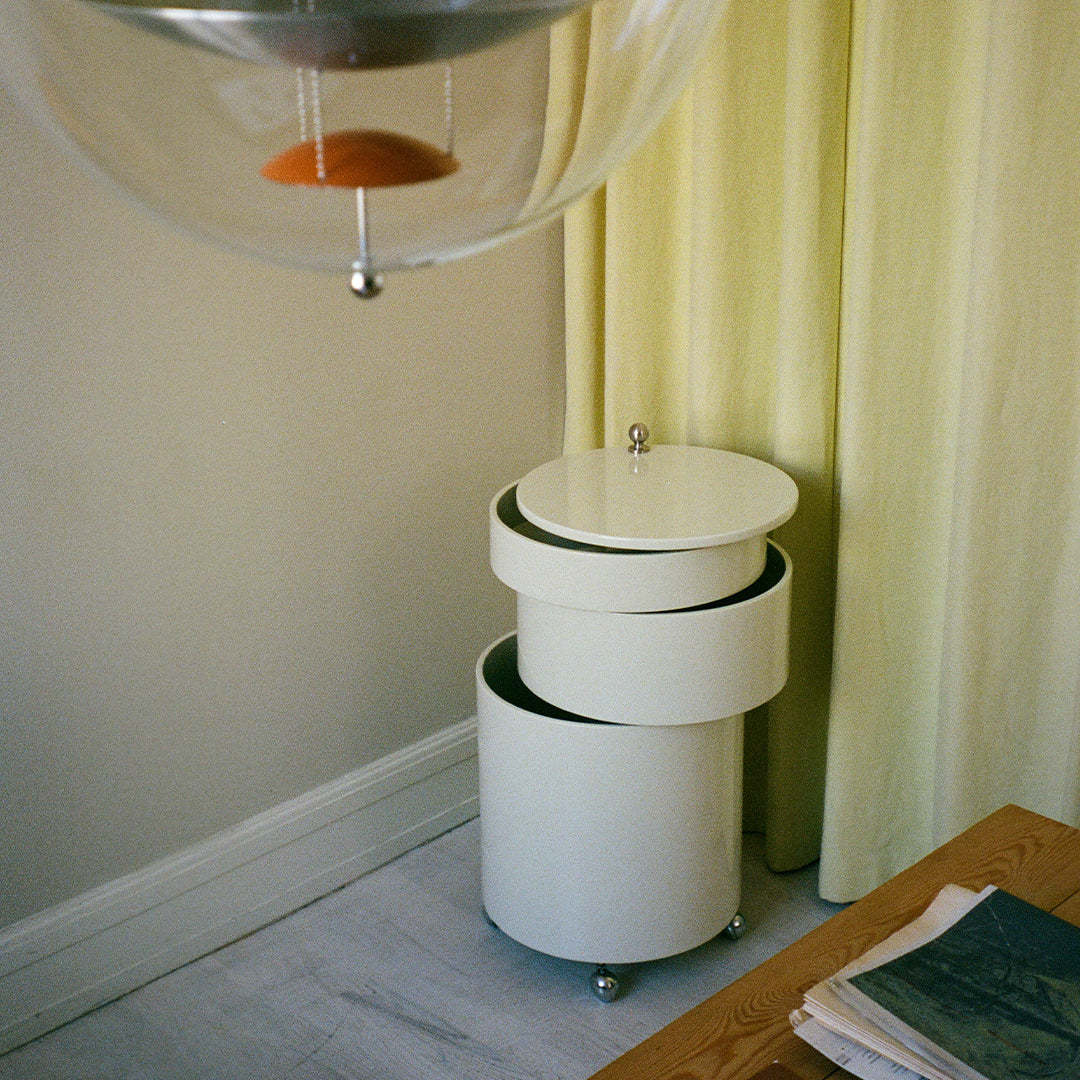 White circular side table with smooth surfaces and minimalistic design, set against soft yellow curtains and light wood.