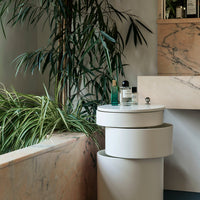 Modern white cylindrical side tables with a smooth finish, set against natural green foliage and marble textures.