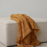 Textured mustard blanket with white stripes draped over a beige ottoman. Perfect for adding warmth and style.