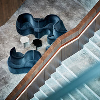 Curved dark blue velvet sofa with black and white accent tables on textured gray flooring. Modern aesthetic.