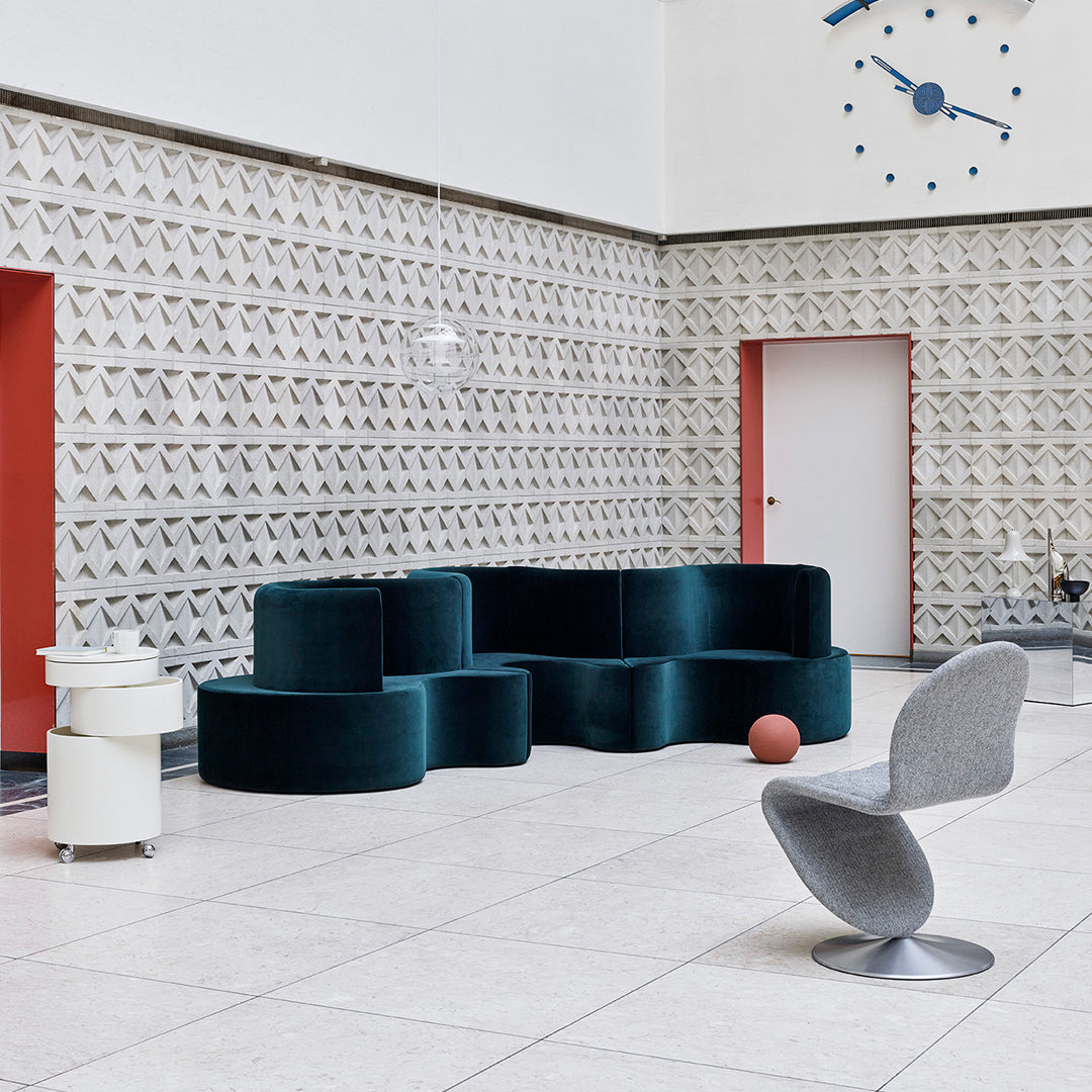 Modern teal velvet sofa and gray sculptural chair against a neutral patterned backdrop with red accents.