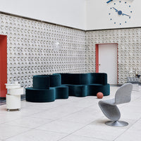 Modern teal velvet sofa and gray sculptural chair against a neutral patterned backdrop with red accents.