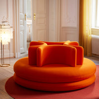 Bright orange circular sofa with unique armrest shapes, set on a red rug in a sunlit room.
