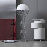 White globe lamp with a spiral silver base next to white cylindrical table and a maroon folder on a gray floor.