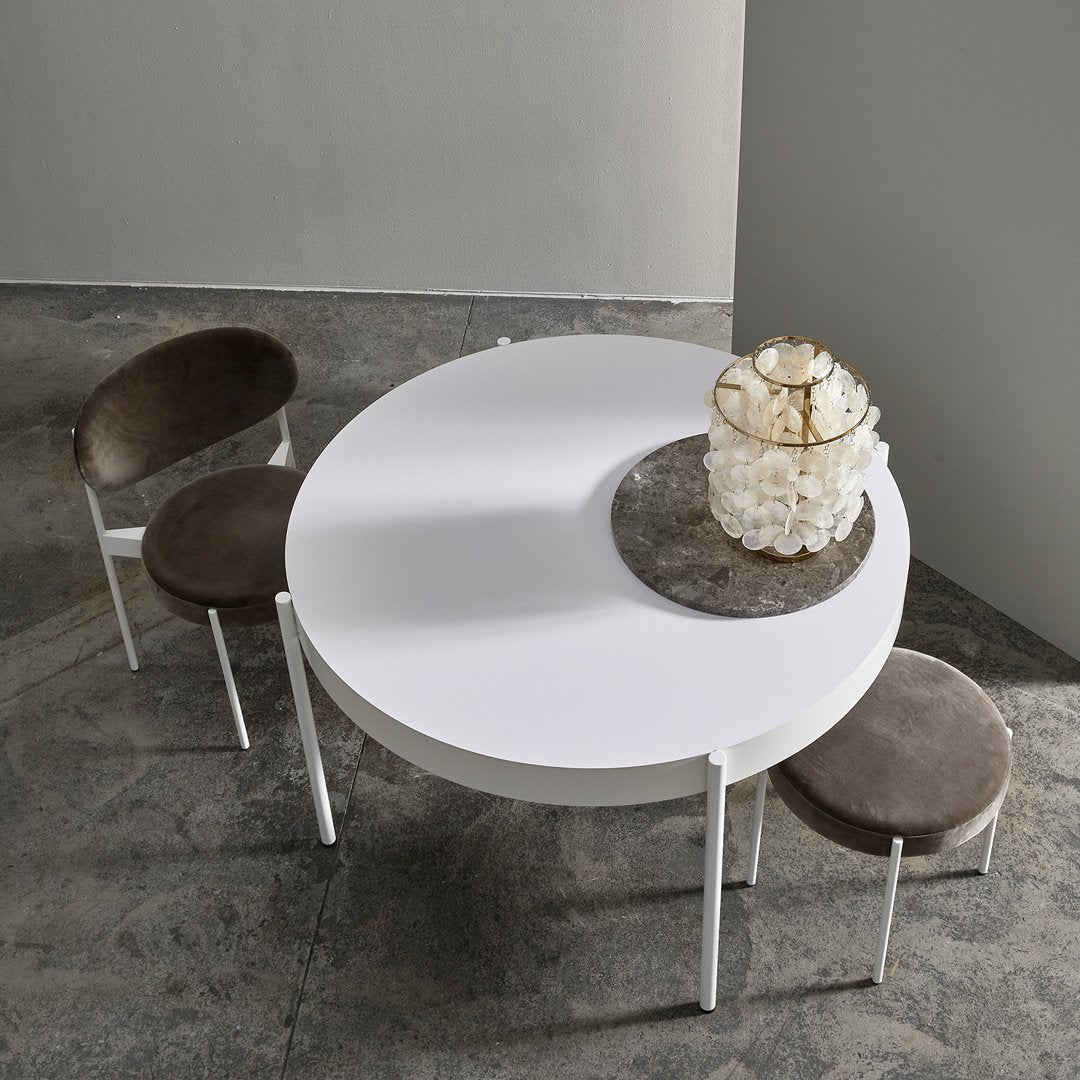 Round white dining table with gray velvet chairs, minimalist design, soft colors, and smooth textures.