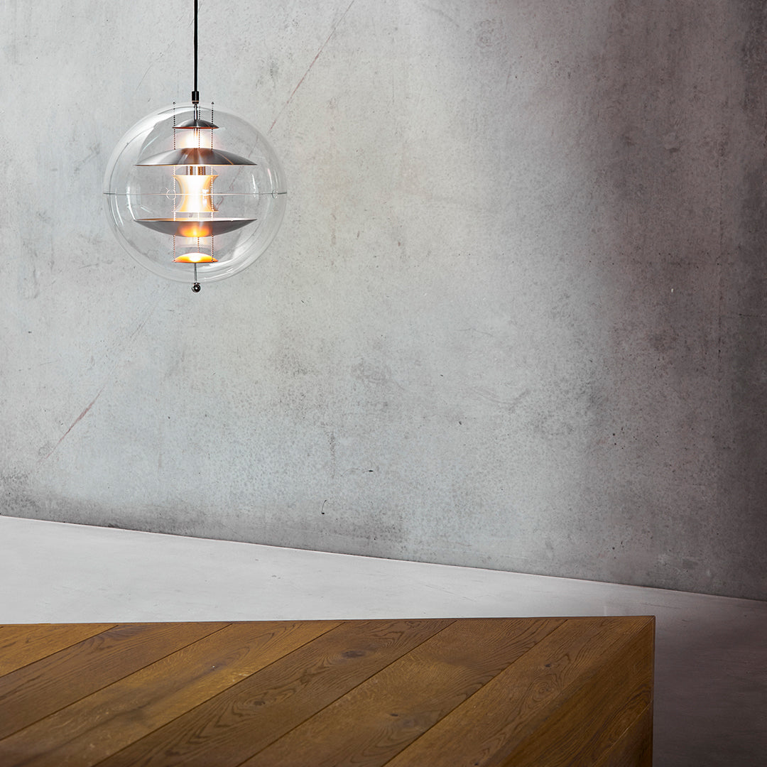 Clear glass pendant light with warm lighting, suspended over a textured gray wall and a rustic wooden table.
