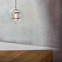 Clear glass pendant light with warm lighting, suspended over a textured gray wall and a rustic wooden table.