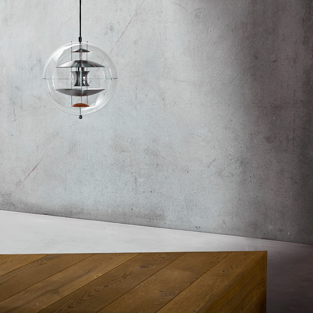 Clear glass pendant light with a metallic inner detail, suspended over a warm brown wooden table against a gray wall.
