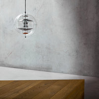 Clear glass pendant light with a metallic inner detail, suspended over a warm brown wooden table against a gray wall.