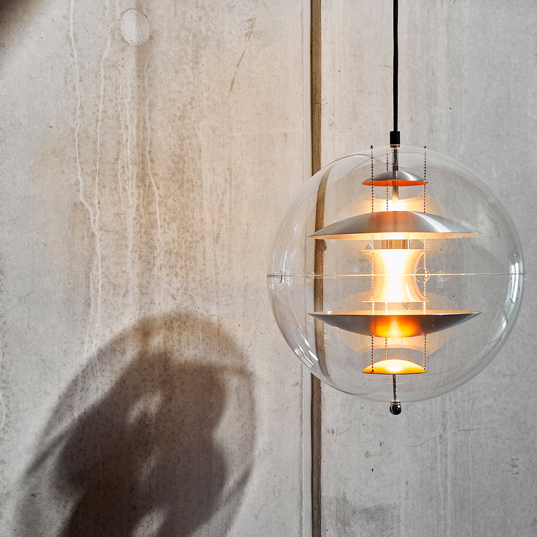 Clear glass pendant light with warm orange accents and layered white patterns, casting soft shadows on a concrete background.