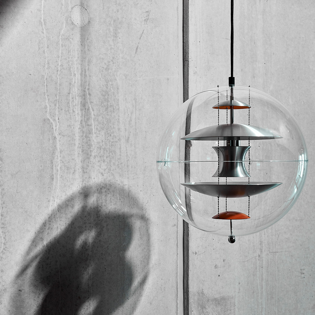 Modern glass pendant light with layered silver and orange accents, casting dramatic shadows on a concrete wall.