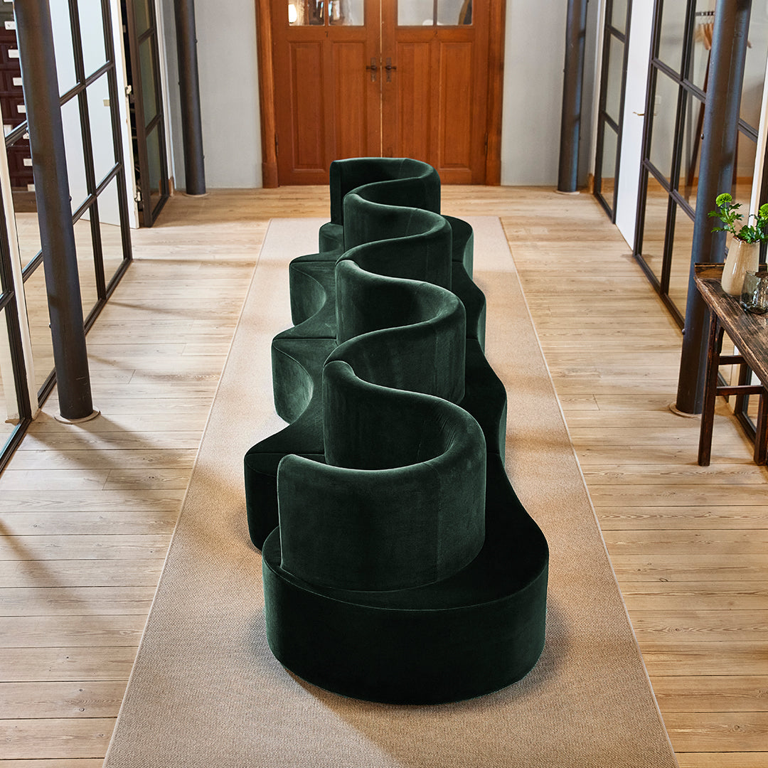 Luxurious dark green velvet sofa with a unique, wavy design, positioned on a neutral-toned rug in a stylish interior.