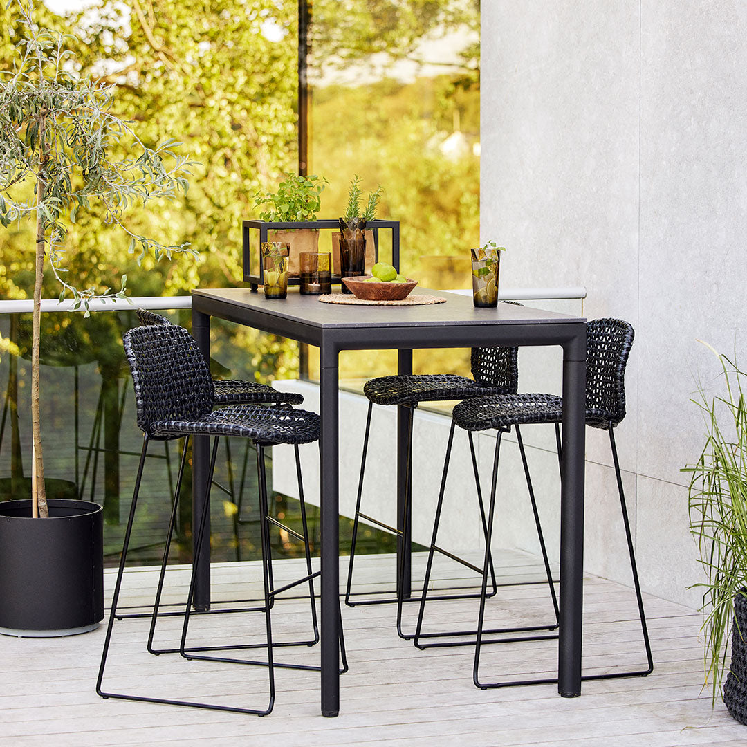 Black outdoor dining table with matching woven bar stools against a backdrop of greenery. Modern and stylish design.