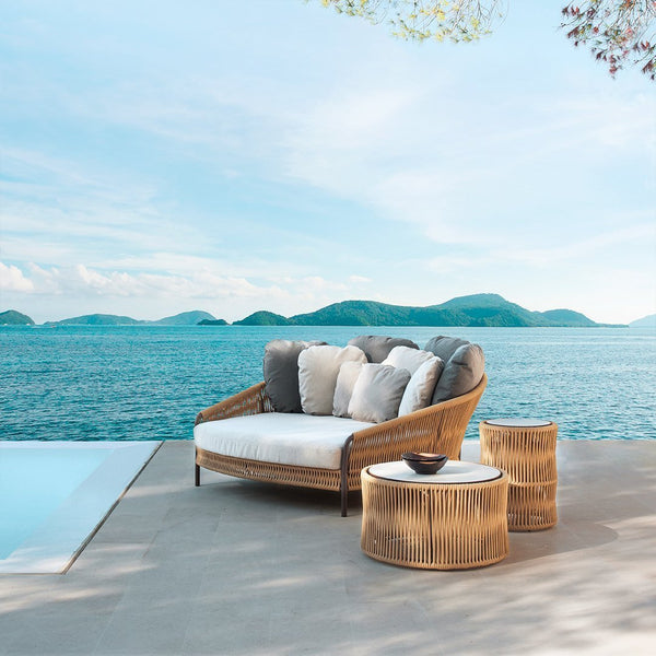 Outdoor sofa with gray and white cushions, paired with natural wicker-style tables against a serene blue ocean backdrop.