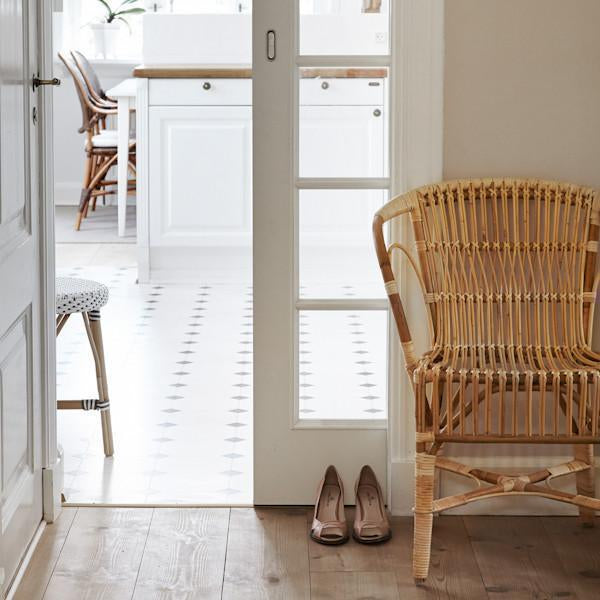 Woven rattan chair in a light beige tone, set against white walls and patterned floor tiles in soft gray.