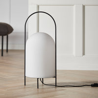 White ribbed lamp in a black arch frame, set on a wooden floor with a neutral background.
